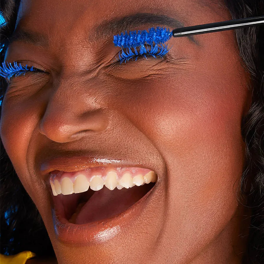 Close-up of a woman laughing with eyes closed as bright blue mascara is applied to her lashes.