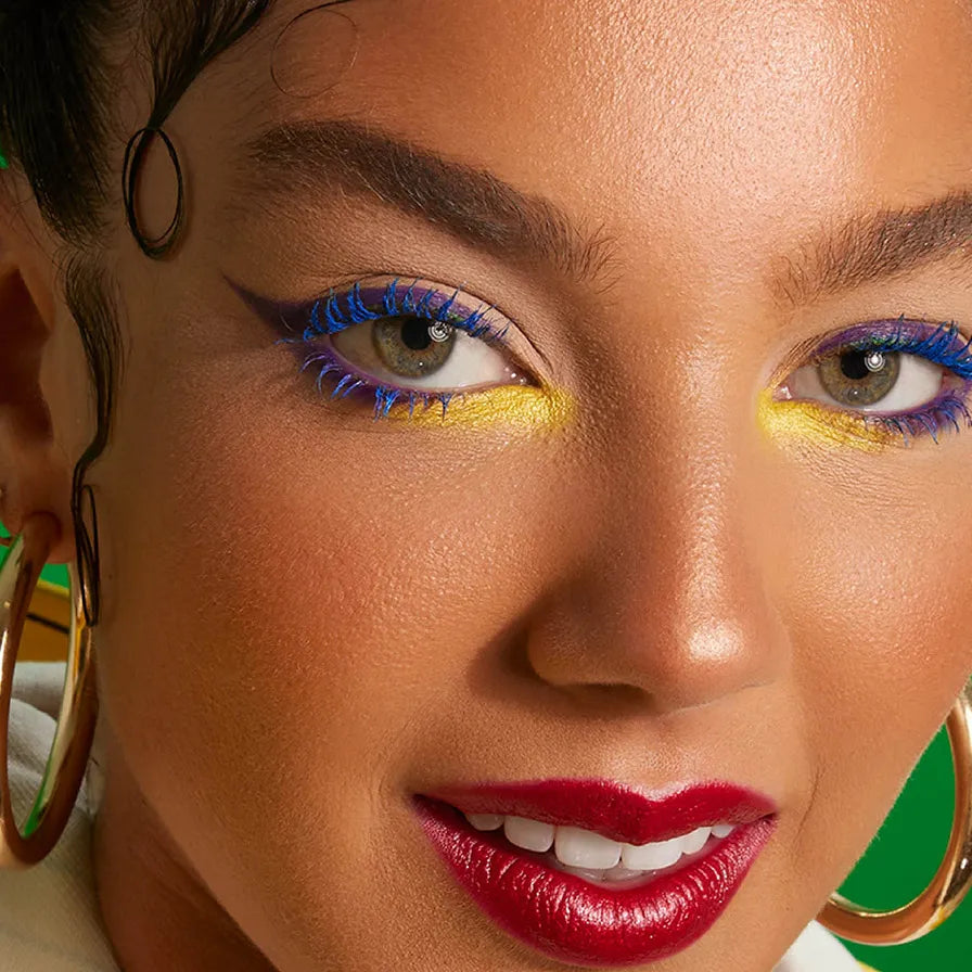  Woman with green eyes wearing bold makeup, featuring electric blue mascara, yellow inner corner shadow, and red glossy lips.