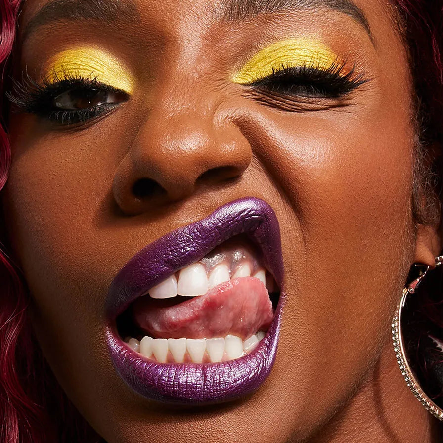  Bold portrait of a woman playfully sticking out her tongue, wearing shimmering purple lipstick and vibrant yellow eyeshadow.
